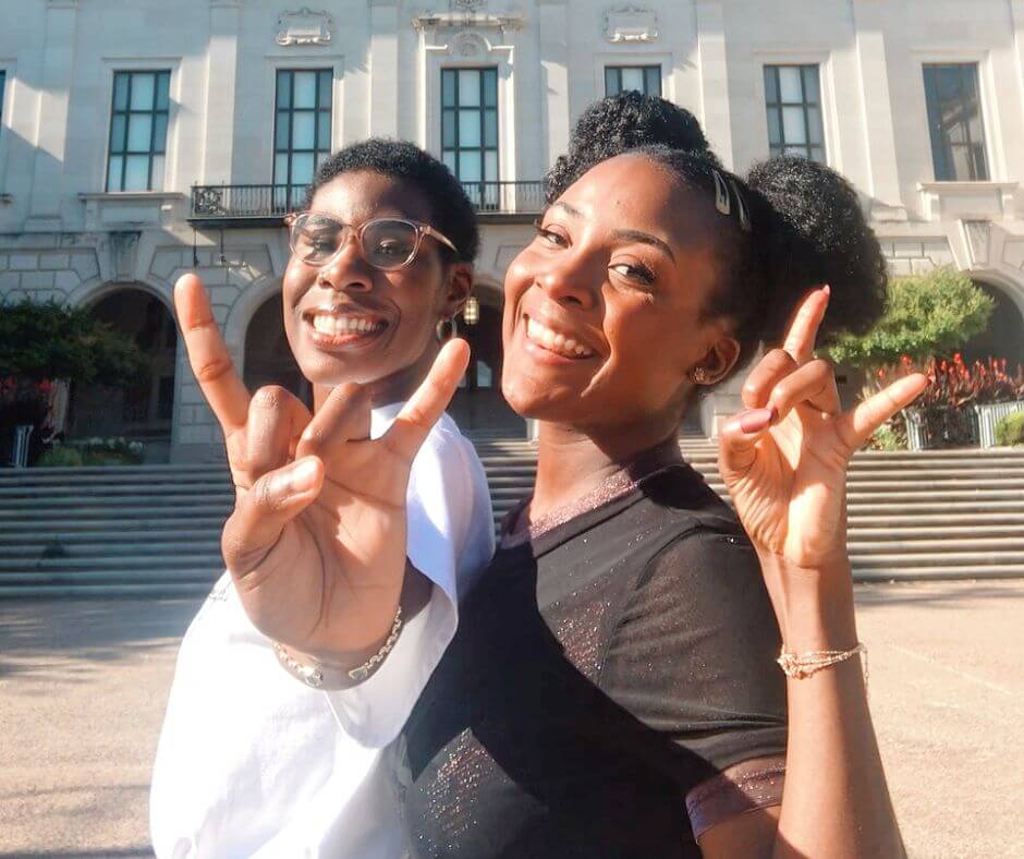 Two female students giving hook 'em horns near the tower on campus