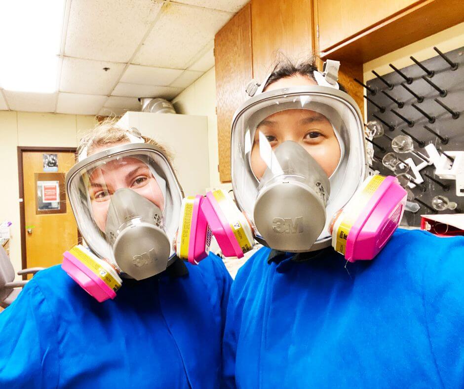 Two female students wearing gas masks while working in a lab